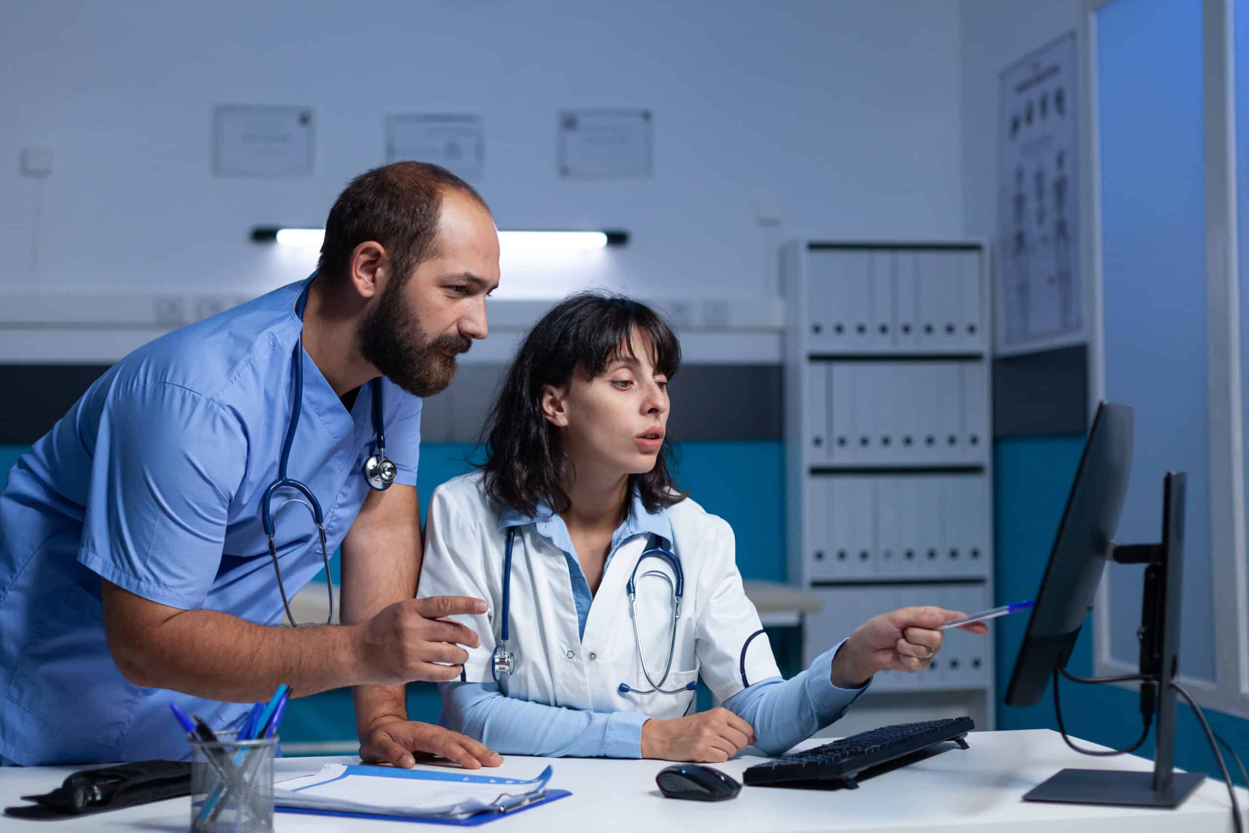 Doctor and nurse pointing at computer for checkup information at night. Medical team of man and woman using monitor display with technology for healthcare system and assistance.